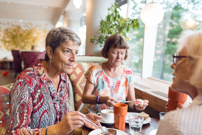 Three women in a cafe sharing customer horror stories while enjoying coffee and dessert in a bright cozy setting.