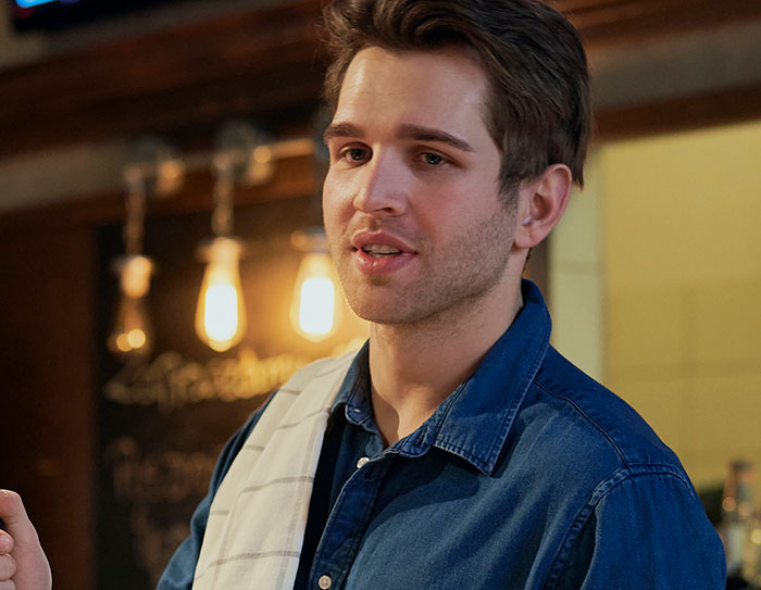 Young male server in a blue shirt sharing customer horror stories in a cozy restaurant setting with warm lighting.