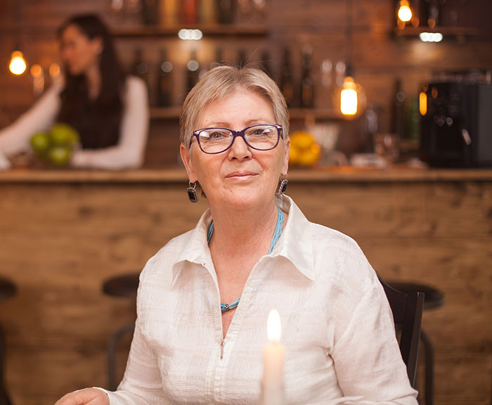 Older woman wearing glasses seated at a restaurant table with a candle, servers sharing customer horror stories atmosphere.