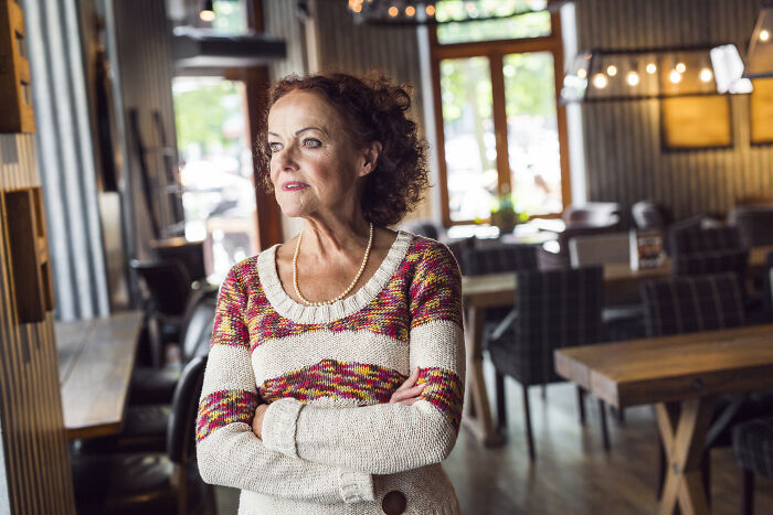 Woman with curly hair in sweater standing inside a quiet restaurant, reflecting on servers sharing customer horror stories.