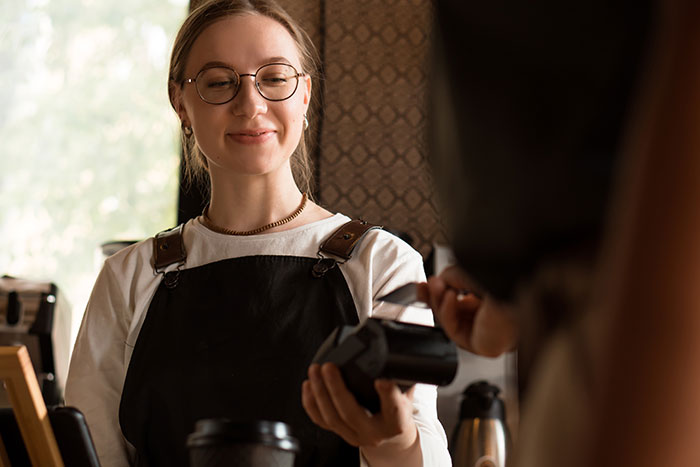 Female server in apron smiling while holding a payment terminal at a cafe with customer horror stories concept.
