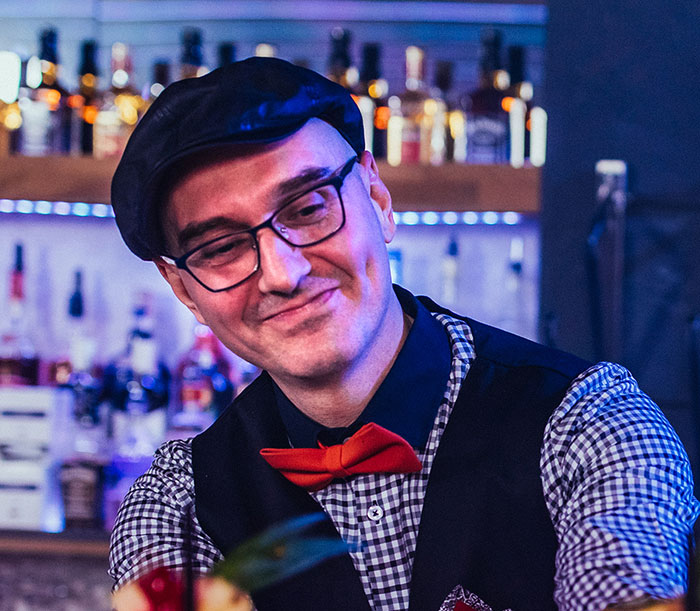 Smiling bartender in a checkered shirt and red bow tie serving drinks, highlighting servers sharing customer horror stories.