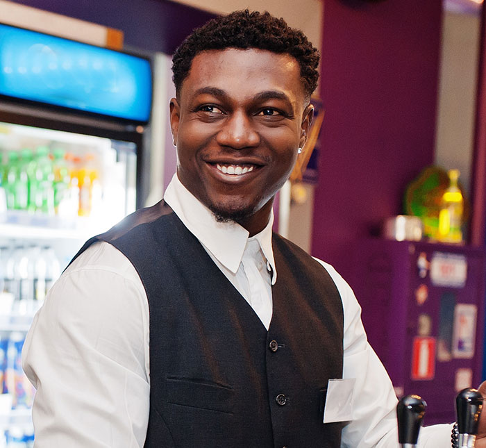 Smiling server in uniform behind bar, representing servers sharing customer horror stories and true nightmares.