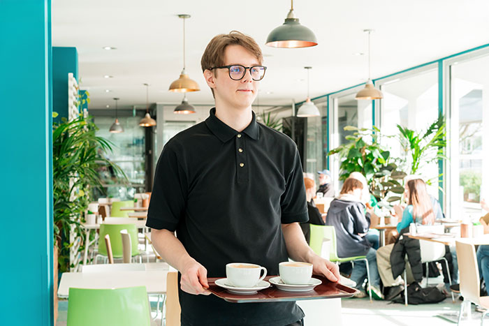 Young server in glasses carrying a tray with coffee cups in a bright cafe, illustrating customer horror stories shared by servers.