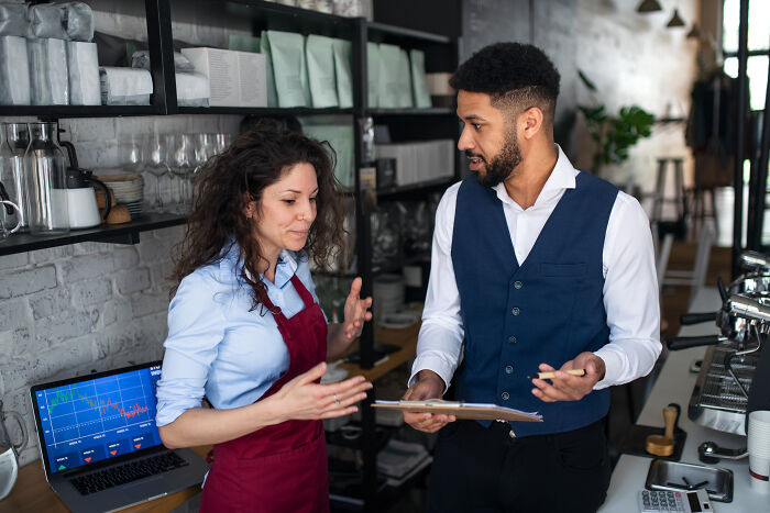A server sharing customer horror stories with a manager in a busy coffee shop setting with equipment and laptop nearby.