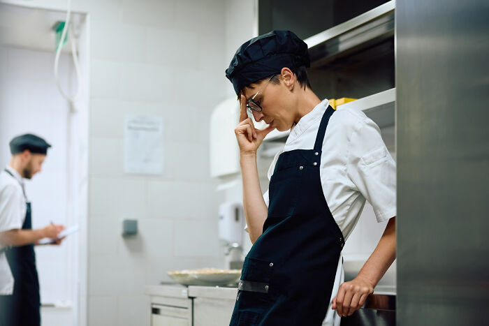 Stressed server in kitchen wearing apron and cap, reflecting on customer horror stories in a busy restaurant environment.