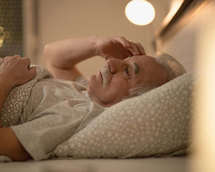 Elderly man lying in bed, holding his forehead, appearing distressed by heartbreaking medical conditions that can't be cured.