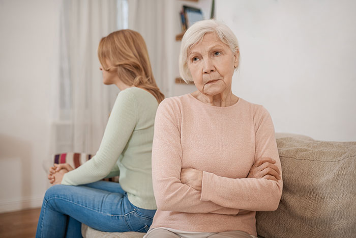 Two women sitting back to back on a couch, looking upset and avoiding conversation amid a tense moment.