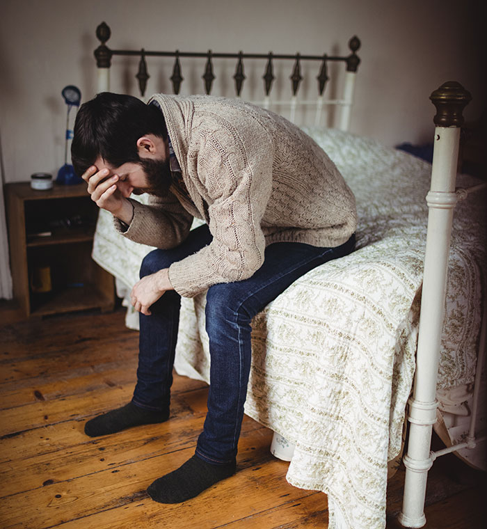 Man sitting on bed, holding head in hand, appearing stressed and deep in thought in a quiet room, lucky times concept.