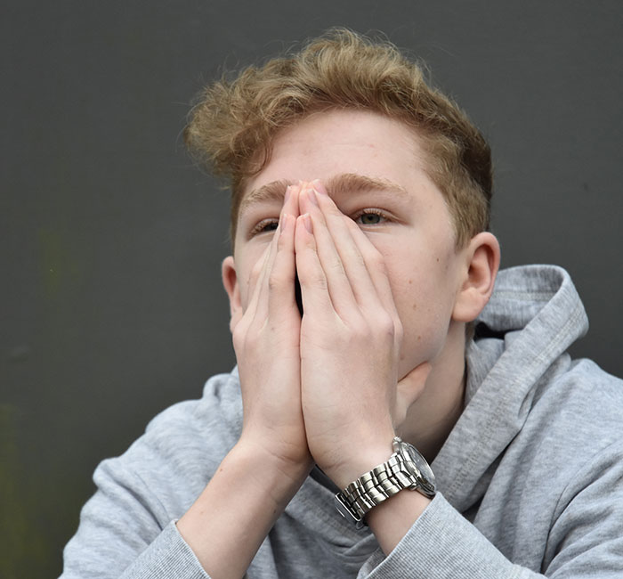 Young person with hands covering mouth, wearing a gray hoodie, reflecting on close calls and intense moments.