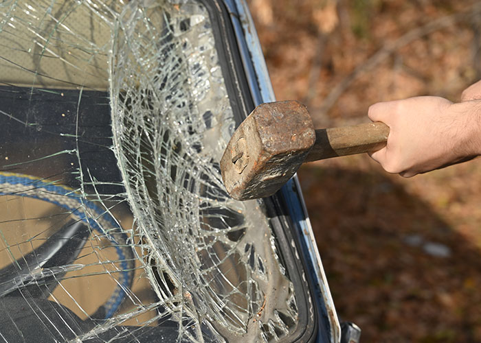Person holding a hammer smashing a car windshield, illustrating people in the wrong place at the wrong time scenario.