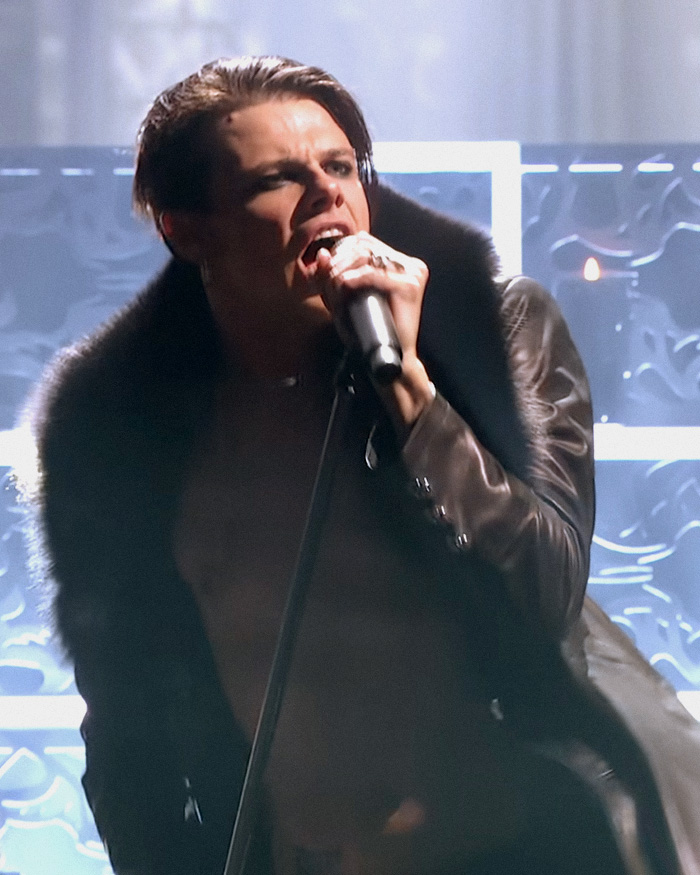 Rock star Yungblud singing passionately on stage wearing a leather jacket with fur collar in a dynamic performance.