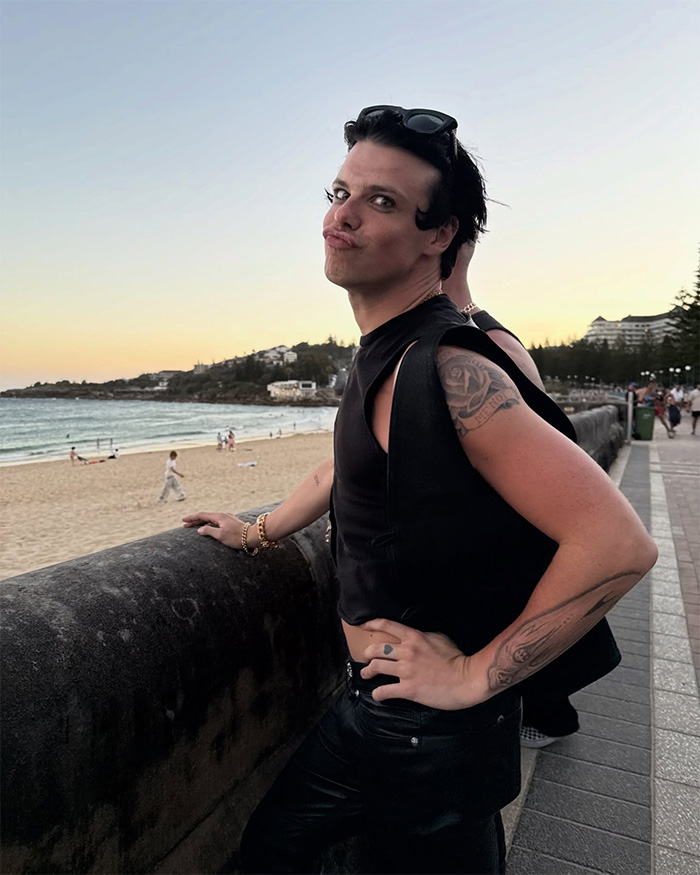 Yungblud posing by the beach at sunset, showing tattoos and wearing a sleeveless black top in casual setting.