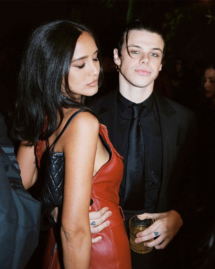 Yungblud dressed in black suit holding a drink, partying with a woman in red dress at a wild Sydney yacht bash.