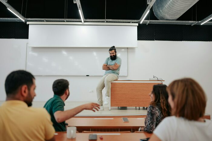 A teacher with a beard and bun sitting on a desk, demonstrating unusual teacher behavior in a classroom setting.