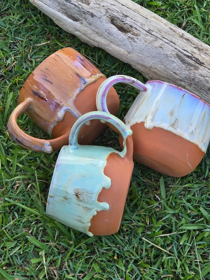 Three handmade ceramic mugs with colorful glaze resting on grass next to a piece of driftwood.