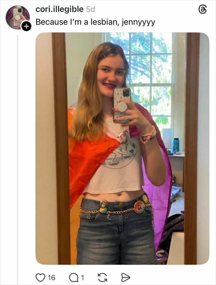 Young woman taking a mirror selfie showing her nails painted, wearing pride flag and casual outfit indoors.