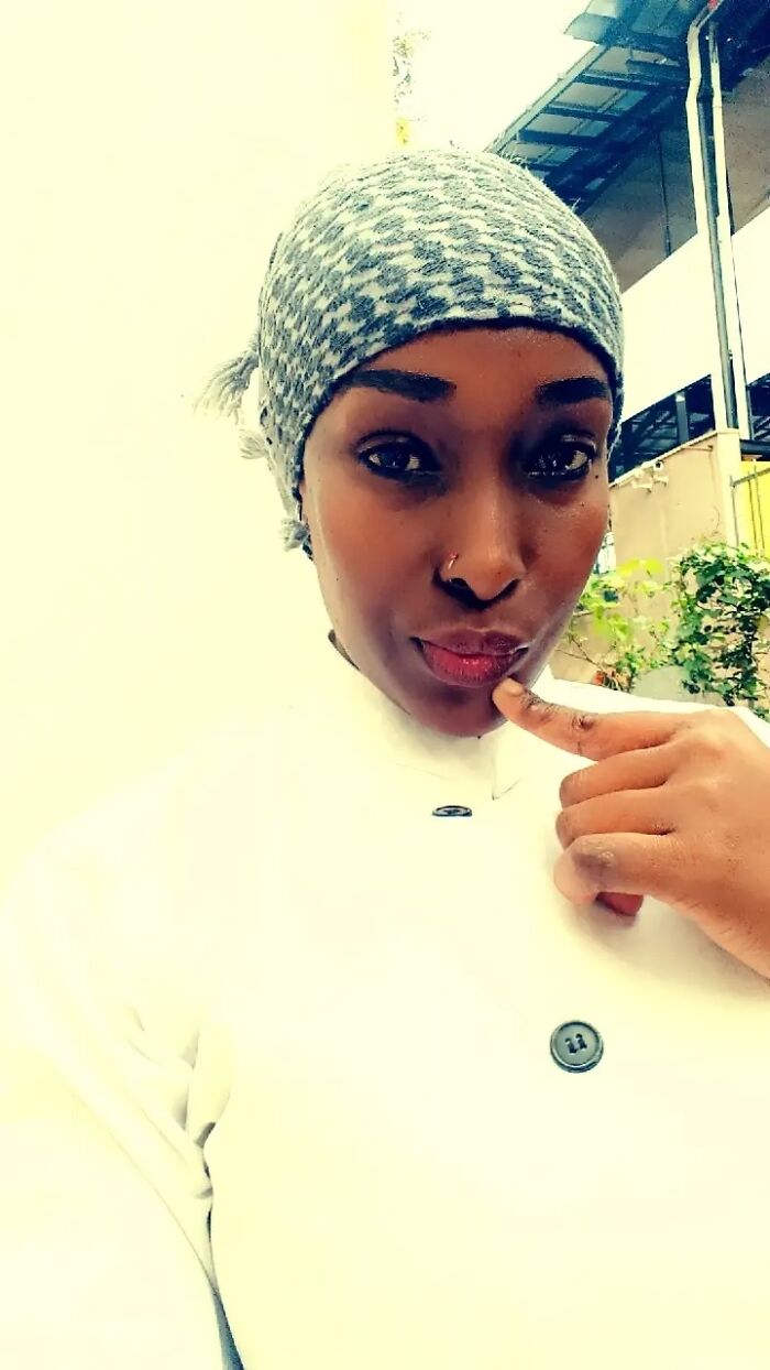 Woman wearing headscarf and white outfit, touching her lips thoughtfully with nails visible in close-up portrait outdoors.