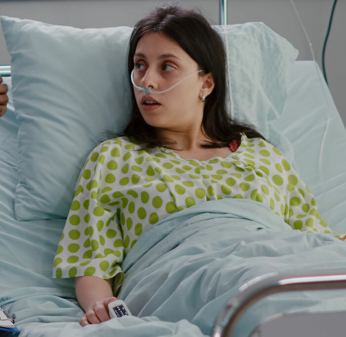 Young woman in hospital bed with oxygen tube, appearing groggy and disoriented after anesthesia recovery.