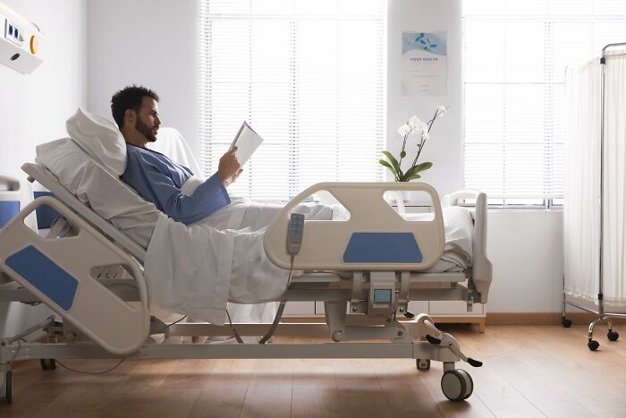 Patient reading a book in a hospital bed recovering after surgery and anesthesia in a bright room.