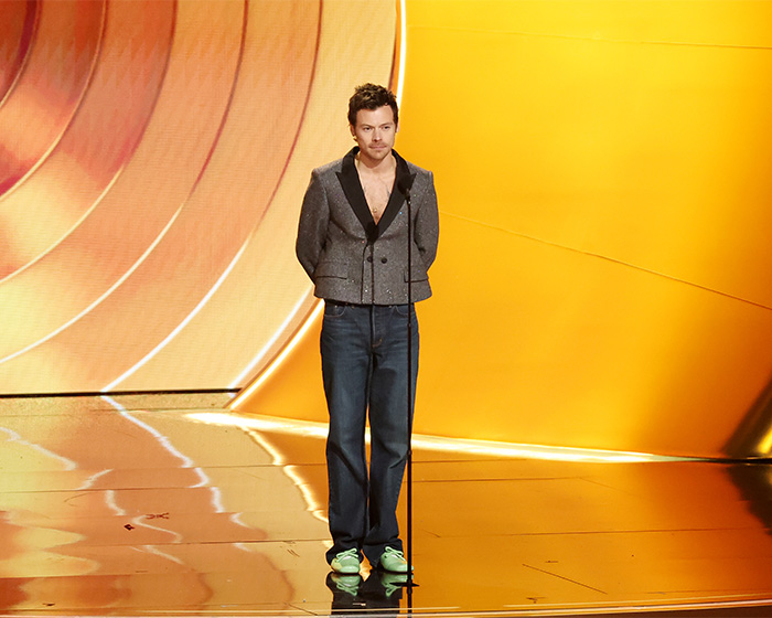 Harry Styles wearing a grey blazer and jeans during on-stage appearance at the 2026 Grammys with orange background.