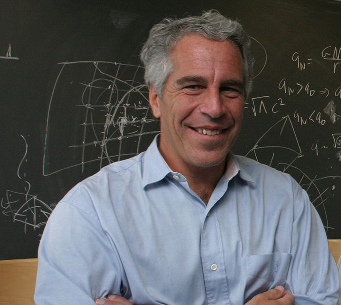 Man with gray hair wearing a light blue shirt standing in front of a blackboard with diagrams, related to Jeffrey Epstein accusations.