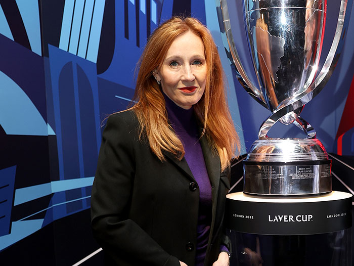 J.K. Rowling with long red hair, wearing a black blazer, standing beside a large silver Laver Cup trophy.