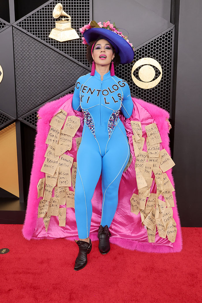 Woman in bright blue bodysuit and floral hat posing with pink fur coat covered in notes on the 2026 Grammys red carpet fashion fails.