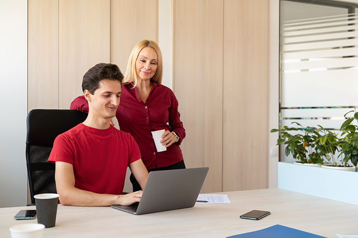 Two coworkers in a modern office setting, discussing something on a laptop during a typical workday scene.