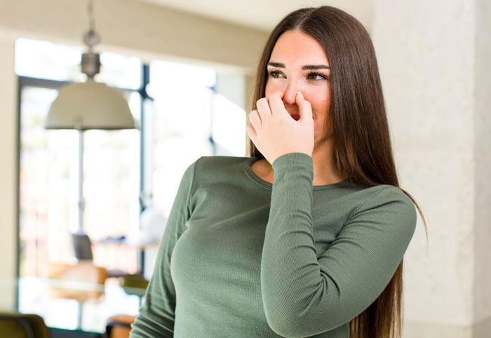 Woman covering her nose indoors, reacting to a stinky smell while standing in a bright home office space.