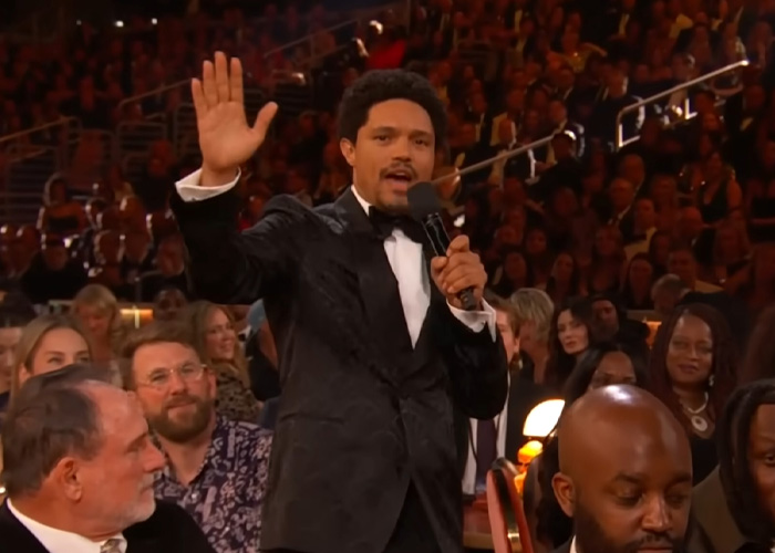 Trevor Noah in a black tuxedo speaking into a microphone at the Grammys, sparking wild theories among the audience.