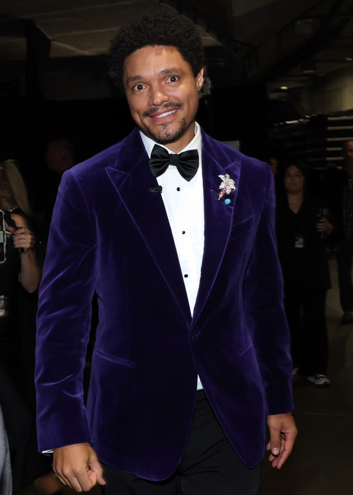 Trevor Noah smiling in a purple velvet jacket and bow tie at a Grammys event backstage with people in the background Trevor Noah smiling in a purple velvet jacket and bow tie at a Grammys event backstage with people in the background