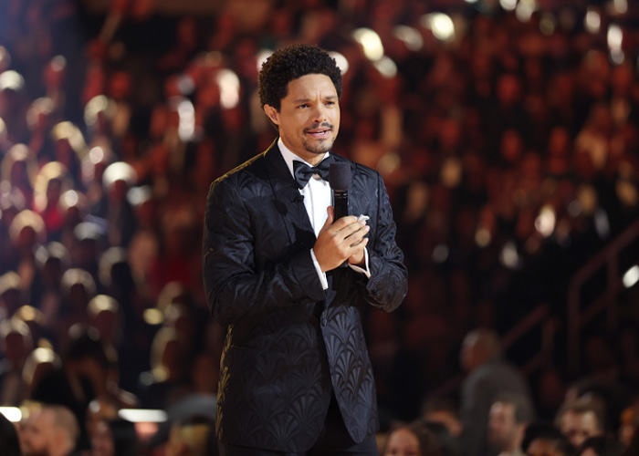 Trevor Noah in a black tuxedo holding a microphone, delivering a bizarre Grammys statement to a large audience.