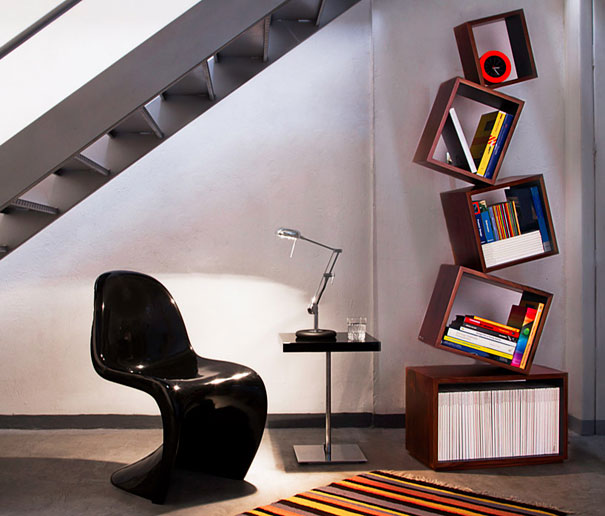 Creative bookshelf design featuring tilted shelves, next to a modern black chair and a small side table under a staircase.