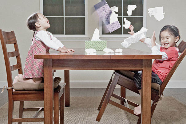Two young daughters laughing and tossing papers at a wooden table in a creative dad&rsquo;s crazy photo setup.