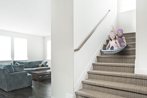 Two daughters wearing winter hats slide down carpeted stairs in a creative dad&rsquo;s playful and crazy photo setup.