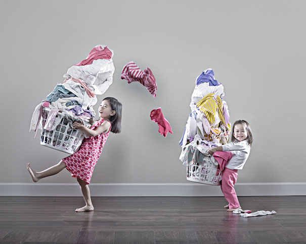Two young daughters hold overflowing laundry baskets with clothes flying, creative dad takes crazy photos capturing the moment.