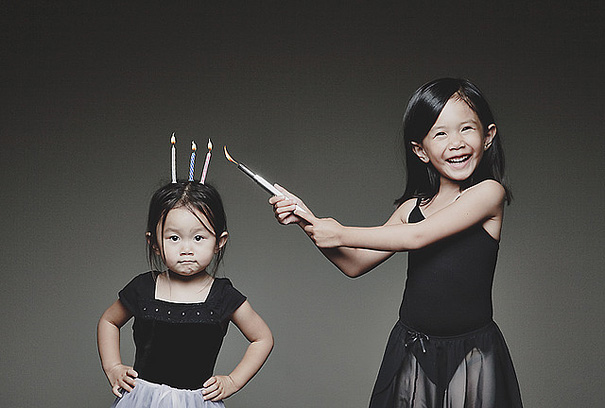 Two young daughters pose creatively with one holding lit candles on the other's head in a playful photo session.