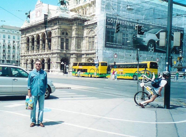 Man posing near a historic building, while a cyclist in the background crashes, creating a perfectly timed photo.