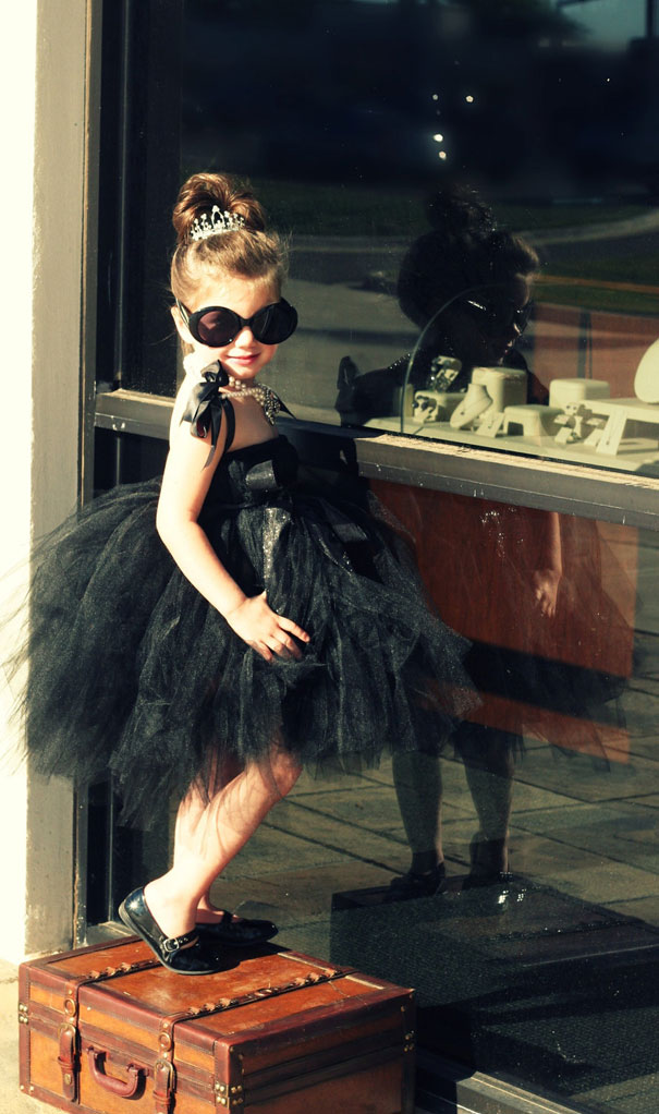 Child in Halloween costume wearing a black dress and sunglasses, posing on a suitcase near a glass window. Child in Halloween costume wearing a black dress and sunglasses, posing on a suitcase near a glass window.