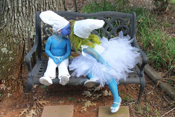 Two kids dressed as Smurfs, featuring white hats and blue body paint, sit on a park bench.