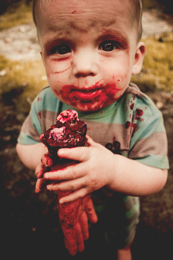 A child in a striped shirt with a Halloween costume, holding a realistic fake hand and covered in fake blood.
