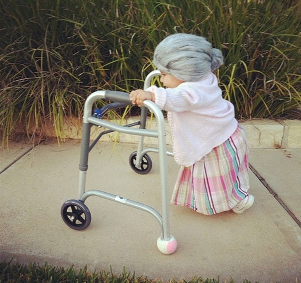 Toddler in a creative Halloween costume, dressed as an elderly lady with a walker, outdoors on a sidewalk.
