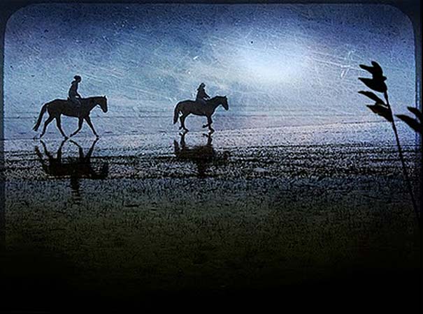 Silhouettes of two riders on horseback reflected on water under a dreamy, blue-toned sky capturing dream imagery. Silhouettes of two riders on horseback reflected on water under a dreamy, blue-toned sky capturing dream imagery.