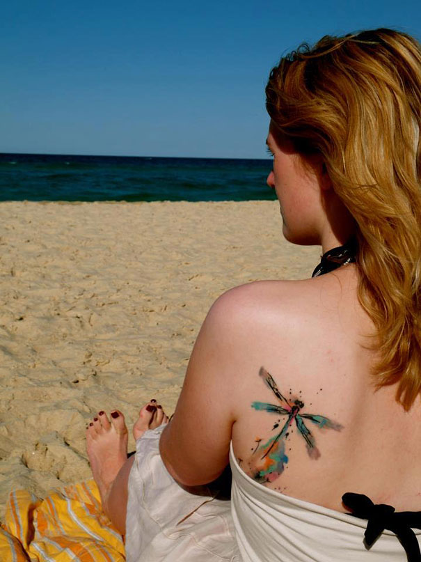 A woman with an artistic watercolor tattoo of a dragonfly on her shoulder, sitting on a beach. A woman with an artistic watercolor tattoo of a dragonfly on her shoulder, sitting on a beach.